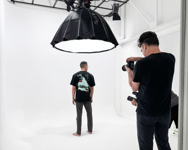 A photographer captures a model wearing a graphic t-shirt in a well-lit studio with a plain white background.