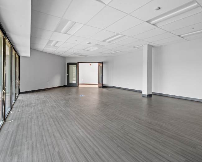 The image shows a spacious, empty room with gray flooring and a large glass wall on one side, featuring two doorways at the opposite end.