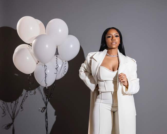 A woman in a white outfit stands next to a cluster of white balloons against a gray background.