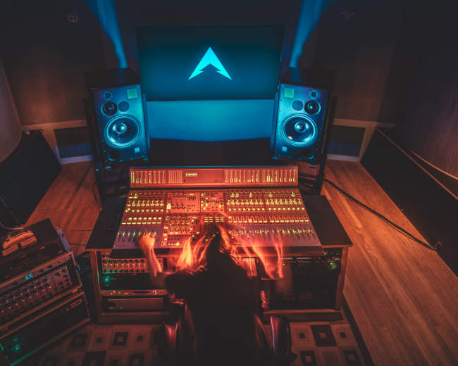 A person is working at a large audio mixing console in a sound studio, surrounded by loudspeakers and a large screen displaying a logo.