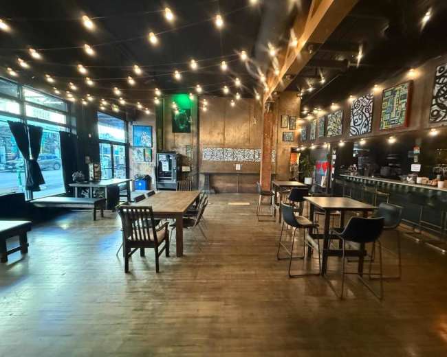 The image shows a spacious café interior with wooden tables, chairs, and a bar area, illuminated by string lights.