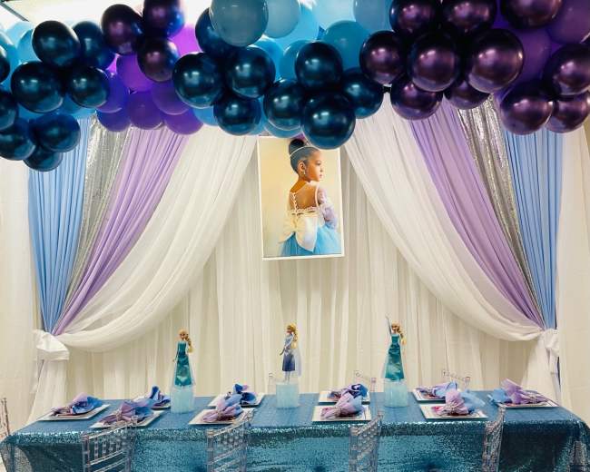 A vibrant party table is decorated with blue, purple, and white balloons, a shimmering tablecloth, and themed table settings featuring character figurines.