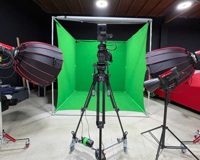 The image shows a studio setup with a camera on a tripod facing a green screen, flanked by two softbox lights.