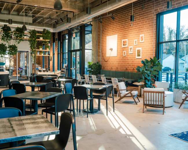 The image shows a modern cafe with large windows, tables, and various chairs arranged around a spacious interior featuring indoor plants and decorative wall art.