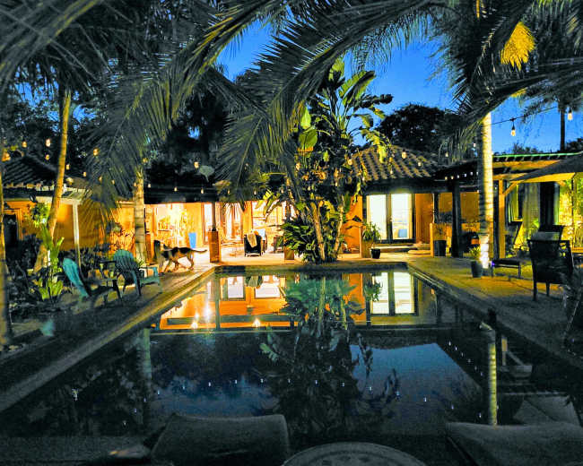 A serene nighttime scene of a swimming pool reflecting the surrounding palm trees and illuminated buildings.