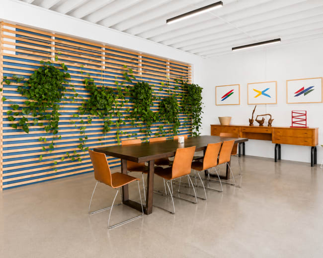 The image depicts a modern dining area featuring a long wooden table surrounded by leather chairs, with a slatted wooden wall and several hanging plants in the background.