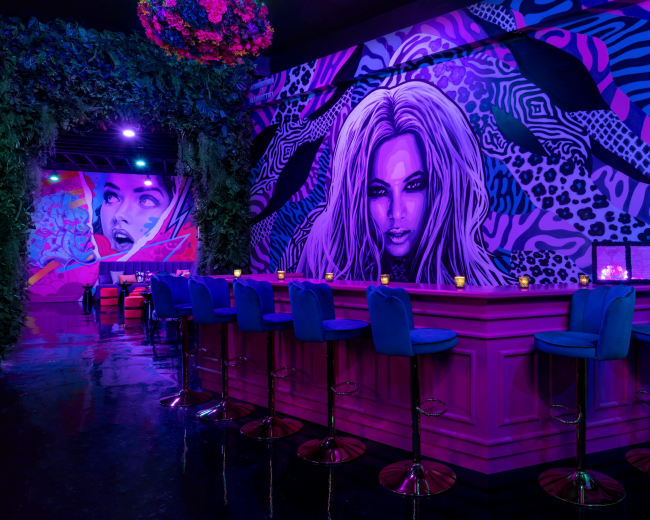 The image features a vibrant bar area with a striking mural of a woman on the wall, surrounded by colorful lighting and modern seating.
