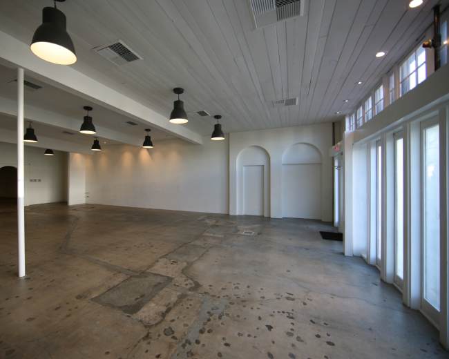 The image shows an empty, spacious room with a concrete floor, white walls, and overhead lighting.
