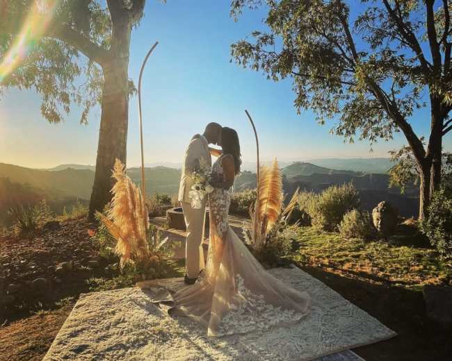 A couple kisses under a tree during their wedding ceremony, with mountains and a clear sky in the background.