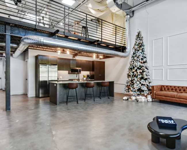 The image shows a modern, industrial-style loft featuring a cozy living area with a brown leather sofa, a minimalist coffee table, a kitchen with stainless steel appliances, and a decorated Christmas tree in the corner.