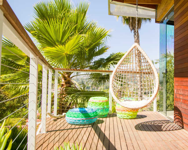 A hanging wicker chair is positioned on a wooden deck beside colorful poufs and surrounded by lush palm trees.