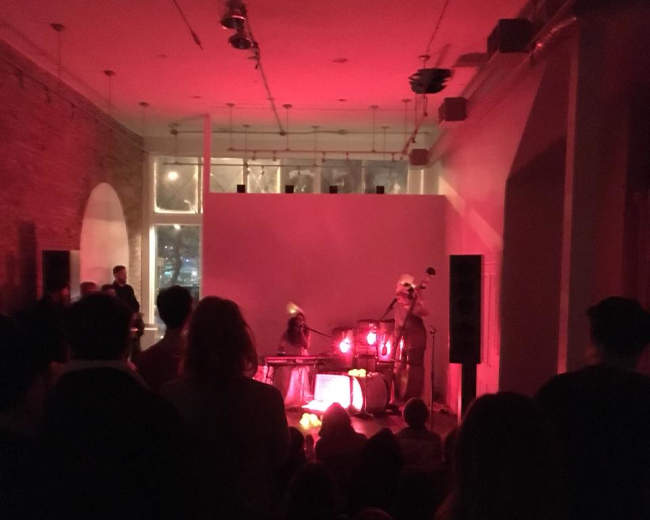 A crowd watches a live performance by musicians on a dimly lit stage, highlighted by red lighting.