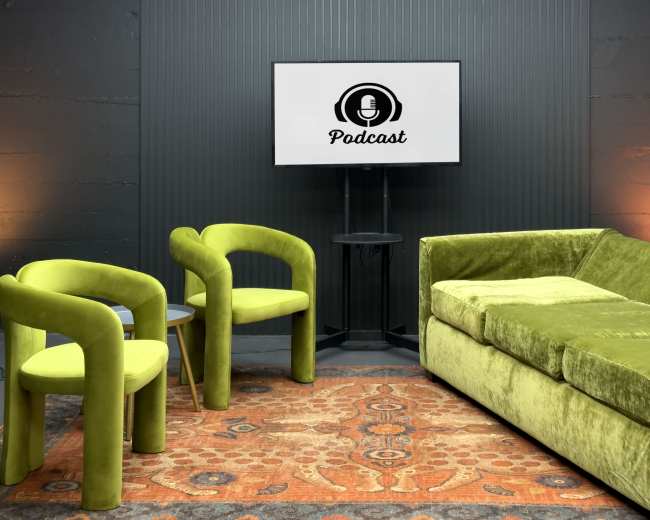 The image shows a podcast studio featuring a green sofa and two matching chairs, with a TV displaying a podcast logo mounted on the wall.