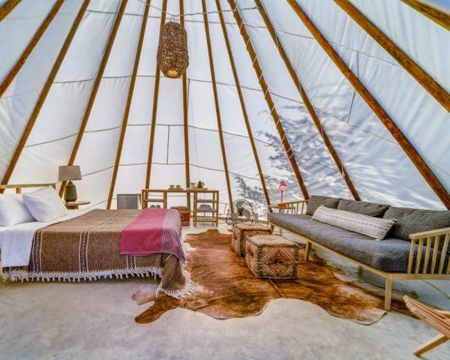 The interior of a glamping tent features a bed, a sofa, and natural wooden furnishings against a backdrop of white canvas walls.