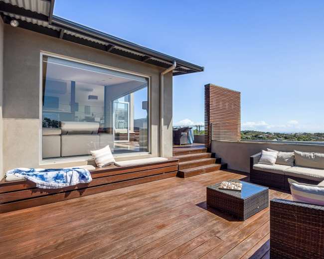 A modern patio features a seating area with cushions, a glass table, and wooden flooring, overlooking a scenic view.