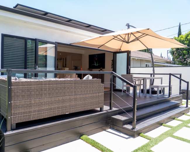 The image shows a modern patio area with a wicker sofa, an umbrella for shade, and steps leading up to an indoor living space.