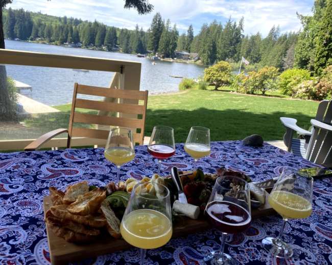 A picnic table adorned with a variety of food and drinks is set near a lake surrounded by trees and a grassy lawn.