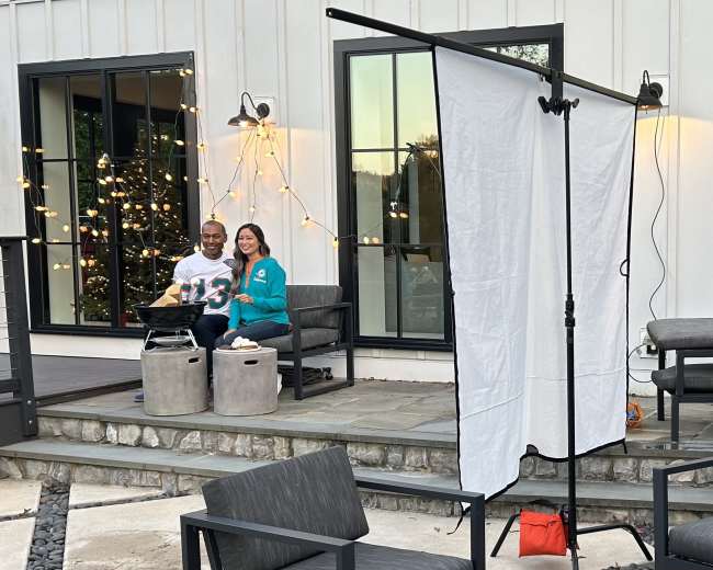 A man and a woman sit on stone stools on a patio, with string lights overhead and a backdrop set up for a photo shoot.