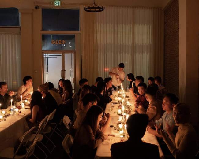 A long table is set with candles and filled with people engaged in conversation during a dinner gathering.