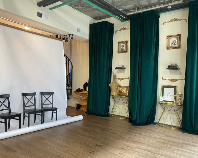 A photography studio featuring three black chairs in front of a white backdrop, flanked by green curtains and gold-framed decorations on the walls.