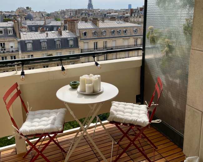 A small balcony features a round white table with two red chairs, overlooking a cityscape that includes the Eiffel Tower in the distance.