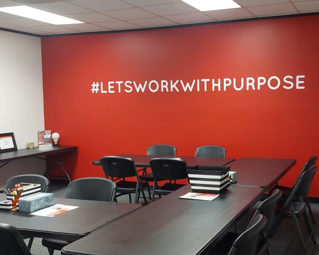 A meeting room with several black chairs and tables arranged under a vibrant red wall featuring the phrase "#LETSWORKWITHPURPOSE."
