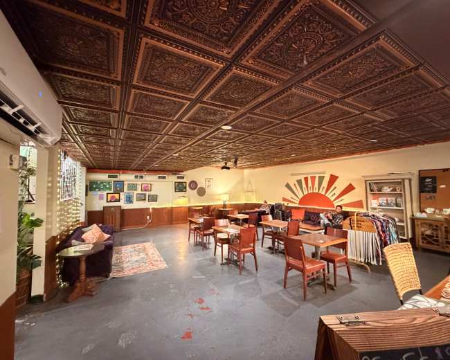 The image shows a spacious café or lounge with a patterned ceiling, various tables and chairs, and colorful artwork displayed on the walls.