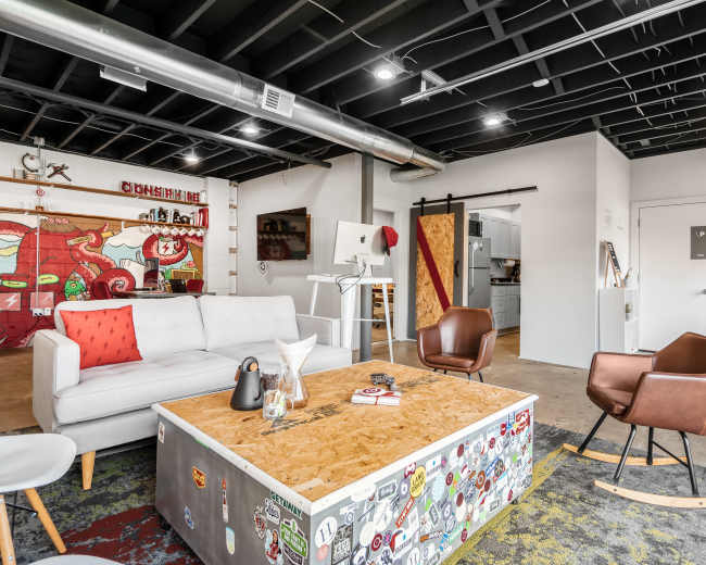 The image shows a modern office space featuring a with a sectional couch, a coffee table adorned with stickers, and kitchen areas in the background.