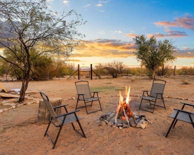 A campfire surrounded by five chairs is set in a desert landscape under a colorful sunset.