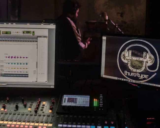 A recording studio setup features two computer monitors displaying audio editing software and a logo, with a musician partially visible in the background.