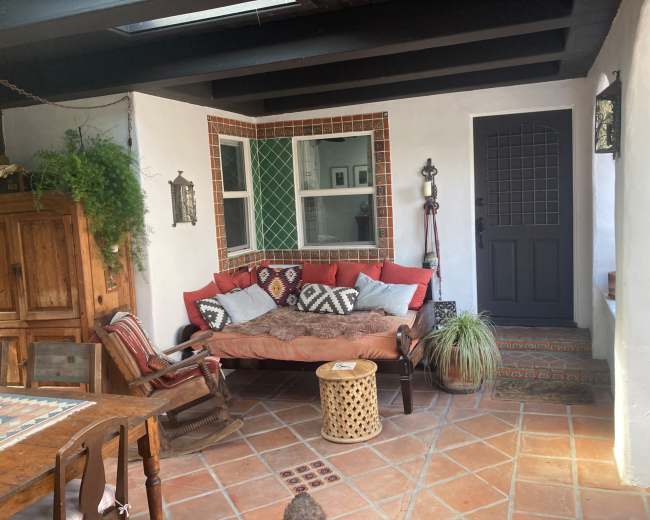 The image shows a cozy courtyard with a rustic wooden sofa adorned with cushions, adjacent to a door and decorated with potted plants and tiled accents.