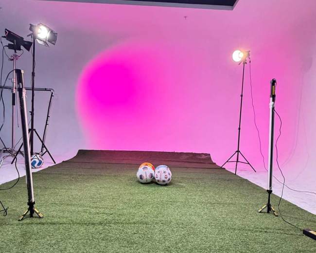 The image shows a studio setup for a photoshoot with artificial grass, two soccer balls, and multiple lighting equipment creating a colorful backdrop.