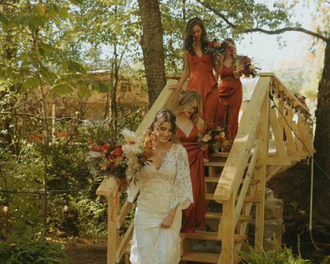 A bride in a white lace dress descends wooden stairs accompanied by three women in red dresses, each holding floral arrangements, surrounded by greenery.