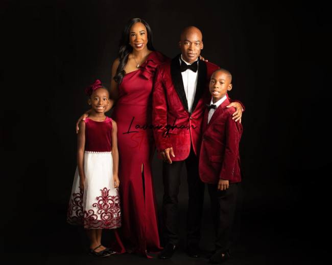 A woman in a red gown and a man in a red tuxedo hold hands with two children, a girl in a red and white dress and a boy in a black tuxedo, against a black background.