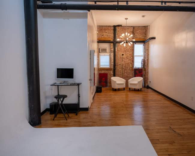 The image shows a spacious, modern room featuring hardwood flooring, exposed brick walls, two white chairs at the far end, and a desk with a computer near the entrance.