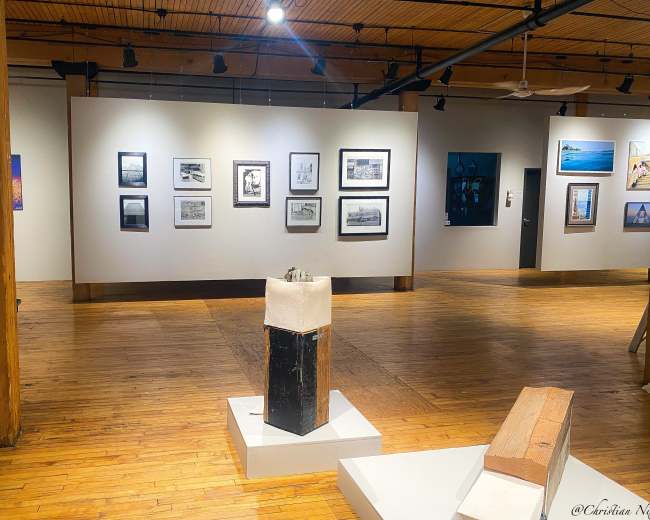 The image shows an art gallery with framed photographs hanging on the walls and a sculpture displayed on a pedestal in the center.