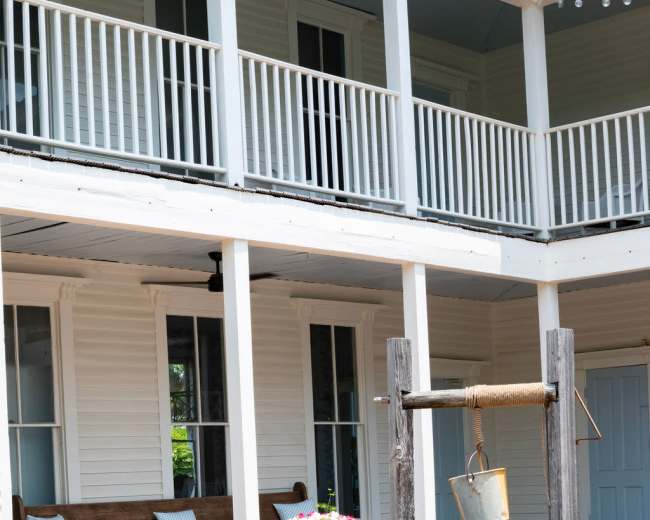 The image shows a stone well in a brick-paved courtyard in front of a two-story wooden house with a covered porch and a bench.