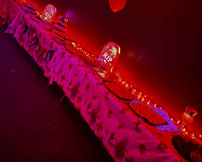 A long table decorated with spider-themed tablecloths, illuminated by red lighting, and featuring items that resemble gravestones with "RIP" inscribed on them.