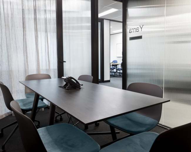 A modern conference room features a rectangular table with a telephone and six teal chairs, surrounded by glass sliding doors with frosted panels.