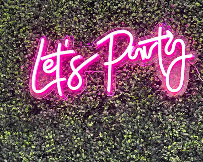 A neon pink sign that says "Let's Party" is mounted on a green wall covered with artificial leaves.
