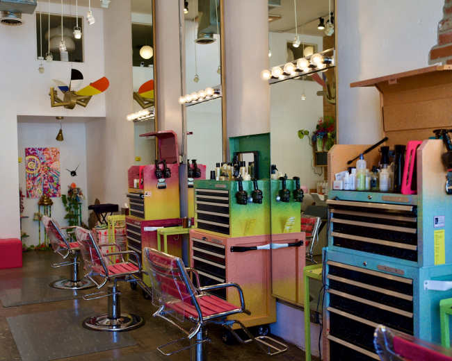 The image shows a brightly colored hair salon featuring multiple styling stations with chairs and storage units against a backdrop of modern decor.