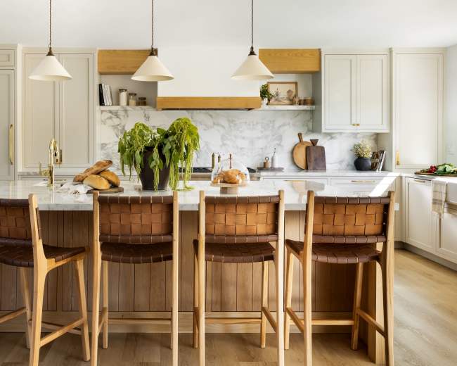 A modern kitchen features an island with six bar stools, a marble countertop, and light fixtures hanging above, with plants and baked goods displayed prominently.
