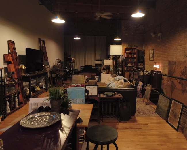 The image shows a spacious, partially furnished loft with wooden flooring, brick walls, and various pieces of furniture, including a couch, bookshelves, and decorative items.