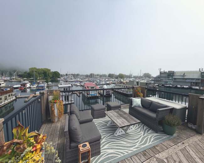 The image shows a balcony area featuring outdoor furniture and a rug, overlooking a marina filled with boats under a cloudy sky.
