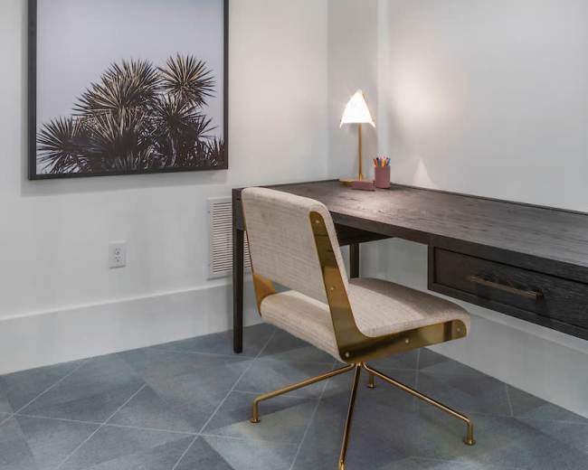 The image shows a modern workspace featuring a wall-mounted framed photograph, a simple desk with a drawer, a stylish chair, and a warm light fixture overhead.