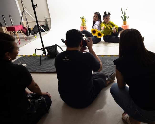 A photographer captures two models posing with sunflowers and plants in a well-lit studio.