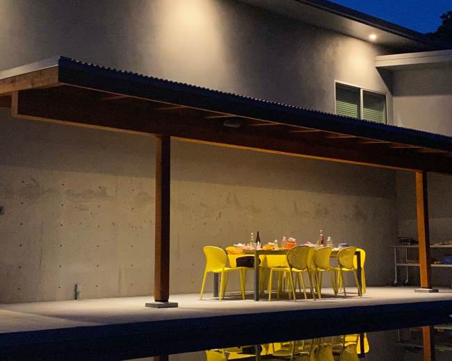 A table with yellow chairs is set under a covered patio next to a pool, illuminated by overhead lights at night.