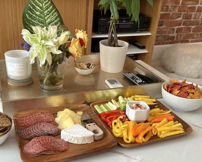 A wooden table displays a charcuterie spread with sliced meats, cheeses, colorful vegetables, and a bowl of dip, alongside a vase of flowers and a potted plant.