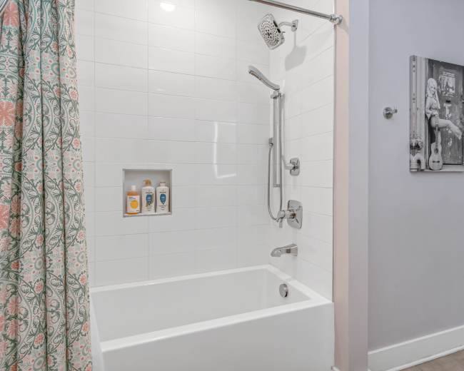 The image shows a bathroom with a bathtub and shower combination, featuring a tiled wall, a showerhead, and a decorative curtain.