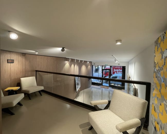 The image shows a spacious interior with two light-colored armchairs placed near a railing overlooking a lower level with large windows displaying a street view.